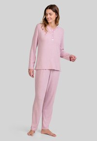 Mujer de pie descalza, vistiendo una camiseta de manga larga de color rosa con canalé y pantalones a juego, frente a un fondo gris claro.