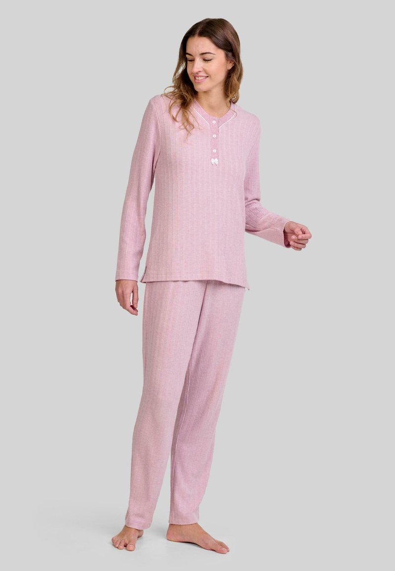 Mujer de pie descalza, vistiendo una camiseta de manga larga de color rosa con canalé y pantalones a juego, frente a un fondo gris claro.