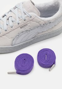 Ljusgrå mockaskor med mjuk foder, blå snören, texturerade detaljer och två rullade lila skosnören med metallspetsar bredvid.