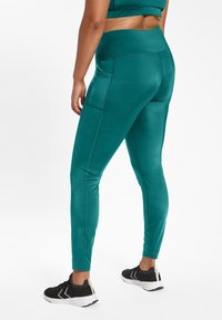 Teal sportleggings met een gladde textuur, hoge tailleband, zijzakken en contrasterende stiksels, gecombineerd met zwarte sportschoenen.
