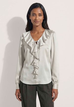 Vrouw met lang donker haar draagt een lichtgrijze blouse met ruches en lange mouwen en donkere broek met fijne strepen, staand tegen een effen achtergrond.