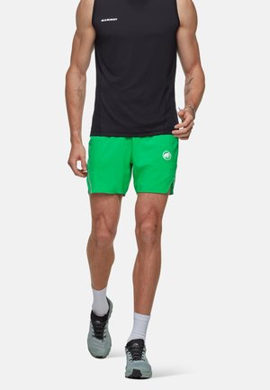 Atletisch persoon draagt een zwart mouwloos topje, felgroene short met logo, witte sokken en grijze hardloopschoenen en loopt naar voren.