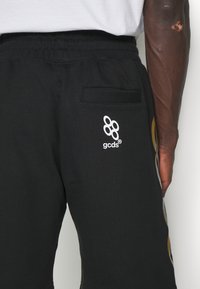 Personne portant un pantalon de jogging noir avec un logo blanc "gcds" sur la poche arrière, une chemise blanche partiellement visible et un bras à la peau foncée.