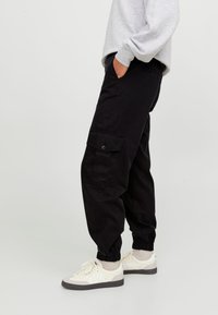 Svarta cargo-byxor med sidfickor, elastiska mudd och en avslappnad passform, tillsammans med vita sneakers, mot en ljus bakgrund.