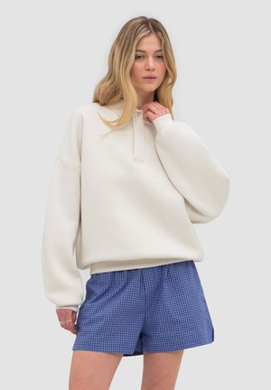 Joven mujer con cabello rubio largo, vistiendo un suéter crema oversize y pantalones cortos azules a cuadros, de pie contra un fondo liso.