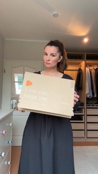 Rjava kartonska škatla s natisnjenim belim besedilom "Ljubim me. Nosim me." in logotipom. Oseba drži škatlo pred dobro organizirano omaro.