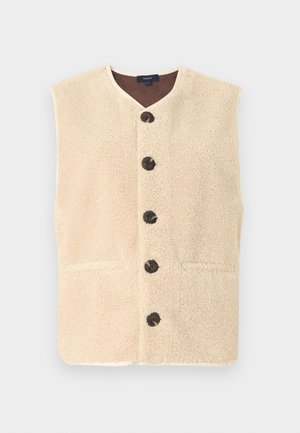 Μπεζ γιλέκο από shearling με μαύρα κουμπιά, καφέ μπλούζα με μακριά μανίκια και καφέ παντελόνια με φαρδιά μπατζάκια. Μαύρα μποτάκια αστραγάλου ολοκληρώνουν το σύνολο.