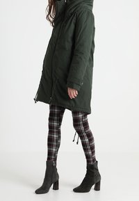 Žena oblečená v tmavě zelené parce, černobílých károvaných legínách a tmavě šedých semišových kotníkových botách na podpatku, stojící na bílém pozadí.