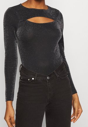 Frau trägt ein funkelndes schwarzes Oberteil mit langen Ärmeln und einem Ausschnitt vorne, kombiniert mit hoch taillierten schwarzen Jeans.