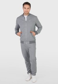 Homme debout, les mains dans les poches, portant une veste grise à fermeture éclair, un pantalon gris assorti et des baskets blanches sur un fond uni.