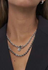 Collana a catena d'argento con due strati: uno con maglie grandi e una chiusura, e l'altro con maglie più piccole alternate e un'etichetta circolare.