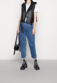 Osoba oblečená v černé kožené vestě přes bílou košili, modrých džínách, černých šněrovacích botách a držící malou černou kabelku.