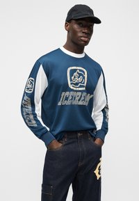 Μακρυμάνικο πουκάμισο από μπλε ναυτικό δίχτυ με λευκές λεπτομέρειες, που φέρει λογότυπο μπροστά και μανίκια με σχέδιο, συνοδευόμενο από σκουρόχρωμο τζιν παντελόνι.