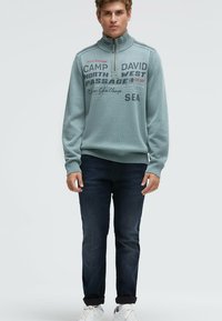 Hellblauer Strickpullover mit hohem Kragen, darauf gedruckter Text in Dunkelgrau und Rot. Kombiniert mit dunkler Jeans und weißen Sneakers.