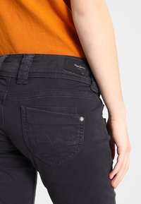 Vaqueros negros con un acabado texturizado, que presentan un diseño distintivo en el bolsillo trasero y una etiqueta de logo. Los vaqueros tienen un corte ajustado y están combinados con una blusa naranja.