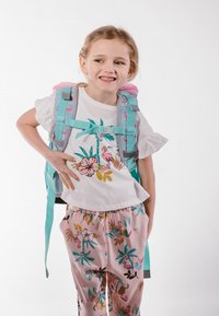Rucksack in Türkis und Pink mit Blumenmustern. Weißes Shirt mit Palm- und Flamingo-Designs, kombiniert mit rosa gemusterten Hosen.
