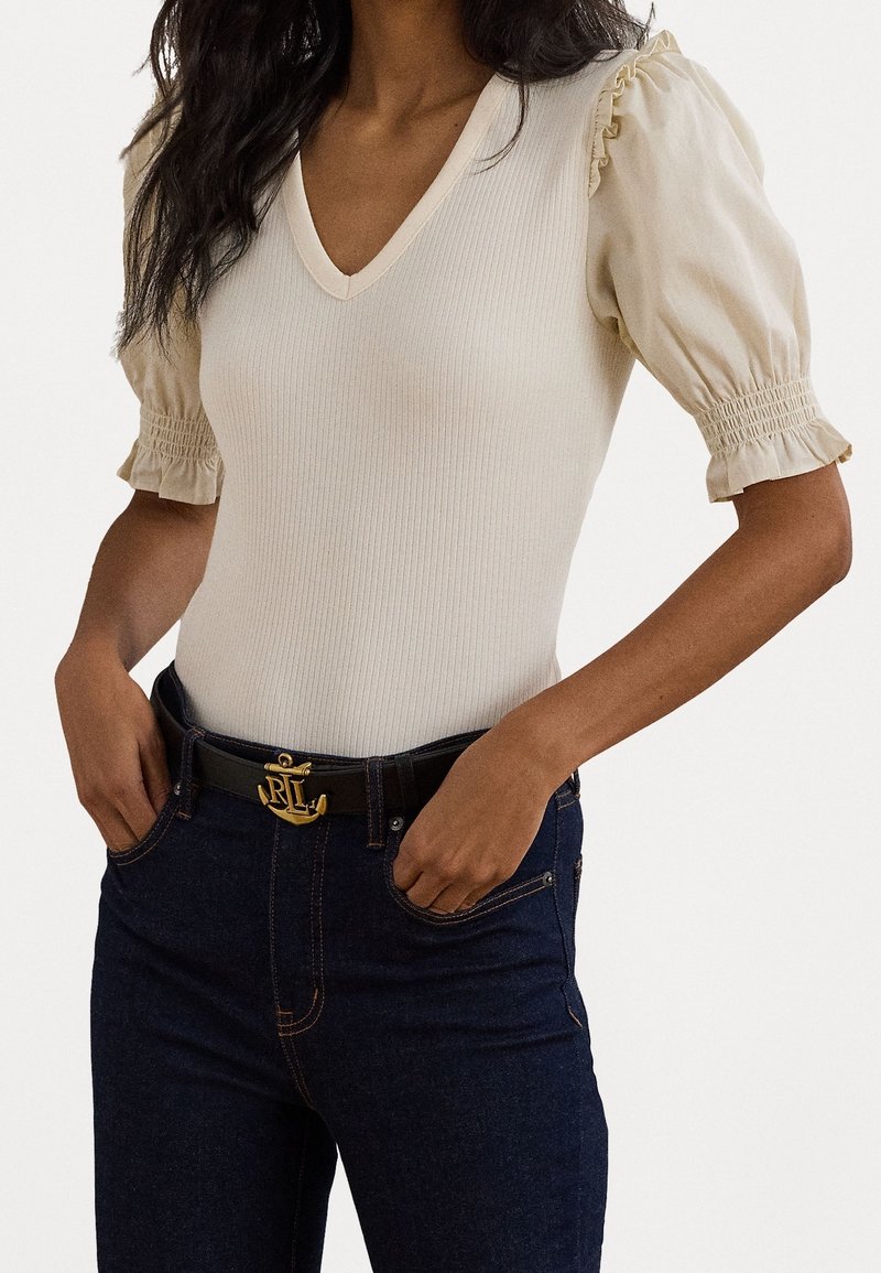 Kvinna iklädd en krämfärgad ribbad V-ringad topp med puffärmar och mörkblå jeans med ett svart bälte som har en guldankarbuckle.