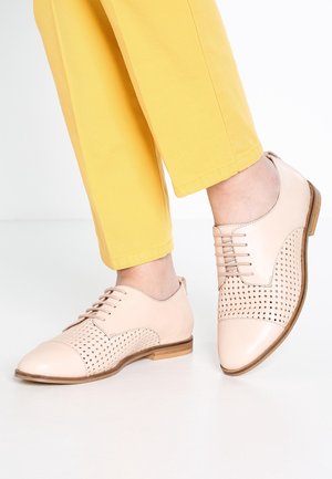 Persoon draagt lichtroze geperforeerde leren oxford schoenen en gele broek, staande op een witte vloer met één voet lichtjes opgetild.