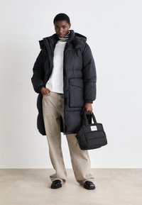 Black puffer coat with a hood, long fit, and front pockets, paired with light beige wide-leg trousers and a small black handbag.