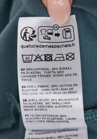 Hoito-ohjeet useilla kielillä. Materiaalikoostumus: 95 % puuvillaa, 5 % elastaania. Valkoinen kangas, jossa on painettu teksti.