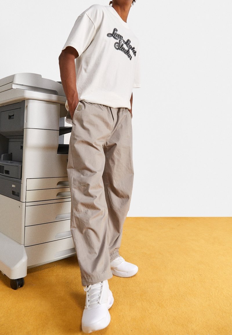 Pantaloni beige larghi, t-shirt bianca con grafica e sneakers bianche. In piedi accanto a una fotocopiatrice grigia su un pavimento giallo.