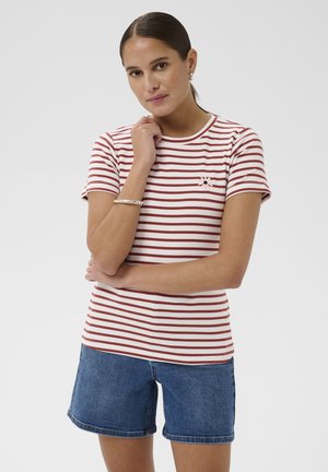 Femme portant un t-shirt à manches courtes rayé rouge et blanc avec un détail fleur, associé à un short en jean bleu, sur fond uni.