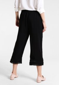 Svarta korta byxor med elastisk midja och spetsdetaljer vid fållen, tillsammans med ljusrosa skor. Slät textur, avslappnad passform.