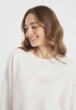 Lachende vrouw met lang bruin haar kijkt omlaag en draagt een witte sweatshirt met roze "WONDER WOMAN" tekst op de voorkant.