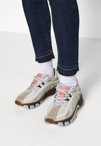 Ljusgrå sneakers med mockadetaljer, rosa och svarta inslag, texturerad sula och en vadderad häl, kombinerade med mörk denim och vita strumpor.