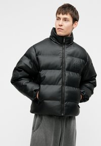 Črna podložena jakna z visokim ovratnikom, gladka tekstura, sprednje zadrgo in elastičnimi manšetami. Ima detajl logotipa na prsih. Topel in moderen dizajn.