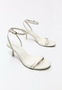 Paire de sandales blanches à talons hauts avec brides à la cheville et petites boucles, avec des talons fins sur fond blanc uni.