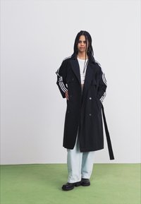 Manteau noir en trench avec un design croisé, des revers larges et des manches longues ornées de rayures blanches. Associé avec des jeans larges de couleur bleu clair.