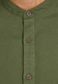 Lähikuva vihreästä paidasta, jossa on pyöreä kaula-aukko ja kolme ruskeaa nappia aseteltuna pystysuoraan etuosan läpikäynnissä.