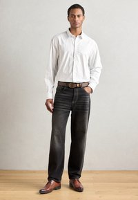 Camicia a maniche lunghe a righe bianche, jeans in denim scuro, cintura in pelle marrone e scarpe eleganti marroni con un motivo decorativo sulla punta. Sfondo piano.
