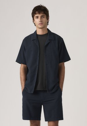 Ung mand står med hænderne i lommerne, iført navyblå tekstureret kortærmet skjorte og matchende shorts over en sort t-shirt.