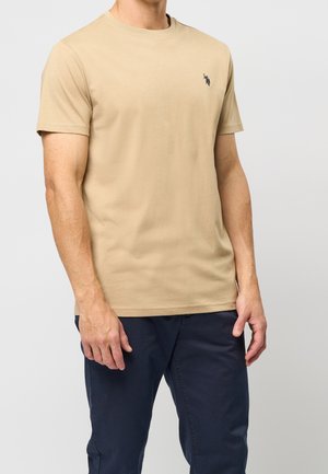 Homme portant un t-shirt beige à manches courtes avec un petit logo noir sur la poitrine et un pantalon bleu marine, debout devant un fond gris uni.