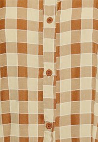 Camisa xadrez castanha e bege com fecho frontal de botões. O tecido possui uma textura suave, apresentando quadrados contrastantes e botões redondos castanhos.