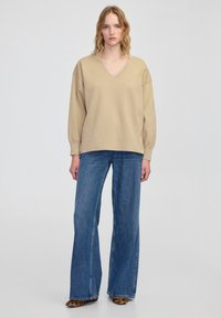 Beżowy sweter w serek o luźnym kroju, z długimi rękawami, zestawiony z szerokimi niebieskimi dżinsami. Dekoracyjne buty na obcasie w wzór zwierzęcy.