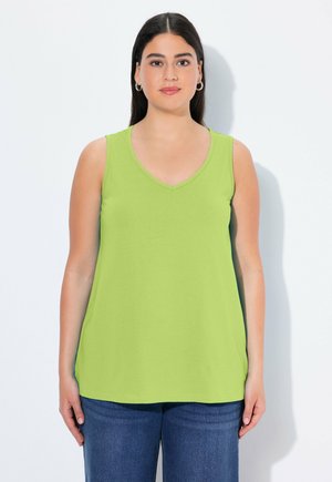 Woman with straight dark hair wearing silver hoop earrings, a sleeveless lime green top, and blue jeans, standing against a white background.