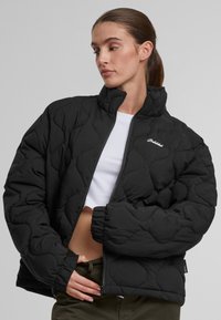 Chaqueta acolchada negra con cuello alto, cierre de cremallera y puños elásticos. Presenta un sutil detalle del logo en el pecho y una textura suave.