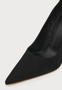 Chaussure à talon haut en daim noir à bout pointu, montrant l'avant et le côté intérieur avec une doublure intérieure noire lisse.