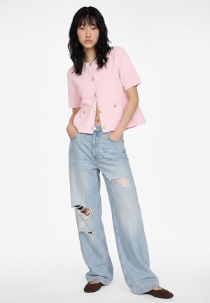 Vrouw draagt een roze kort mouw jasje met knopen, lichtblauwe gescheurde wijd uitlopende jeans en bruine platte schoenen, staand met handen in de zakken.