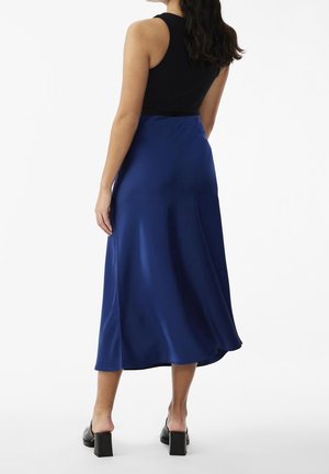 Femme portant un haut noir sans manches, une jupe midi bleue et des mules à talons carrés noires, debout de dos à la caméra.