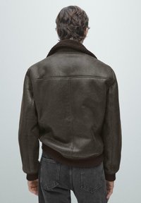 Mand iført mørkebrun shearlingjakke med ribbet manchet og kant, kombineret med falmede sorte jeans, set bagfra mod en ensfarvet baggrund.