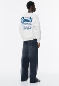 Witte sweatshirt met blauwe grafische tekst en afbeeldingen op de achterkant. Gecombineerd met donkere, wijde jeans en sneakers. De tekst bevat "Peanuts" en activiteiten.