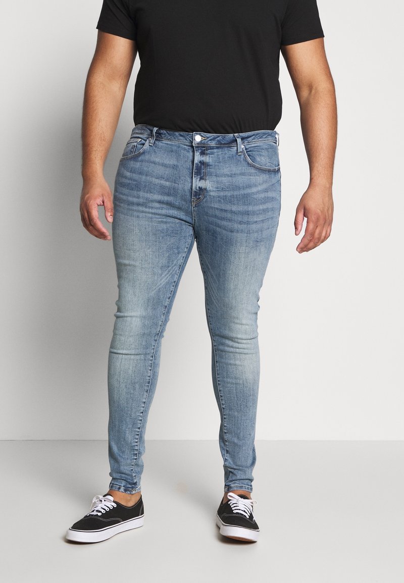 Hombre vestido con jeans ajustados azul desgastados, camiseta negra y zapatillas negras con cordones blancos, de pie frente a un fondo sencillo.