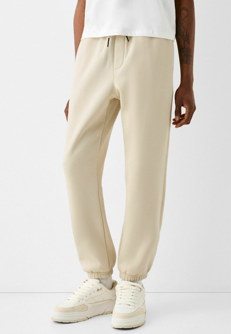 Man som har på sig beige joggerbyxor med elastiska manschetter och vita sneakers, stående mot en helt vit bakgrund.