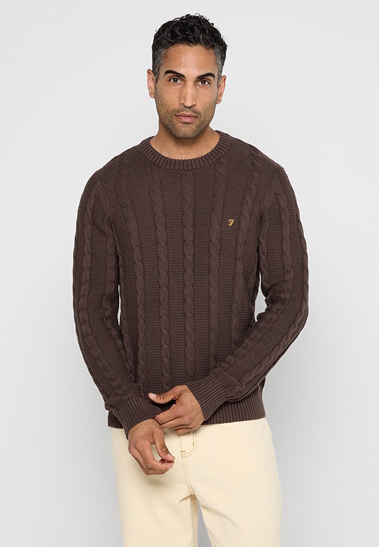 Uomo che indossa un maglione in cashmere marrone scuro a trecce e pantaloni beige chiaro, in piedi con le mani giunte davanti a uno sfondo neutro.