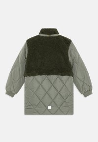 Vadderad jacka med skal i grön quilt och oversized mörkgrön fleecepanel ovanpå, med elastiska ärmslut och tryckknappsstängning.