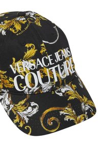 Casquette noire avec des motifs floraux dorés et blancs, arborant un texte blanc audacieux "VERSACE JEANS COUTURE" à l'avant et une visière incurvée.